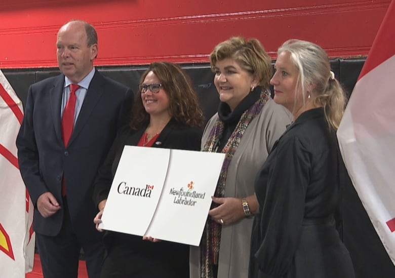 Four people stand in front of Canada and Newfoundland and Labrador flags. The two women in the middle are holding a sign with the federal and provincial government logos on it.