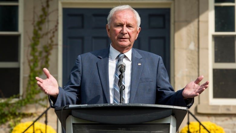 A man in a blue suit stands at a podium outside gesturing with his hands.