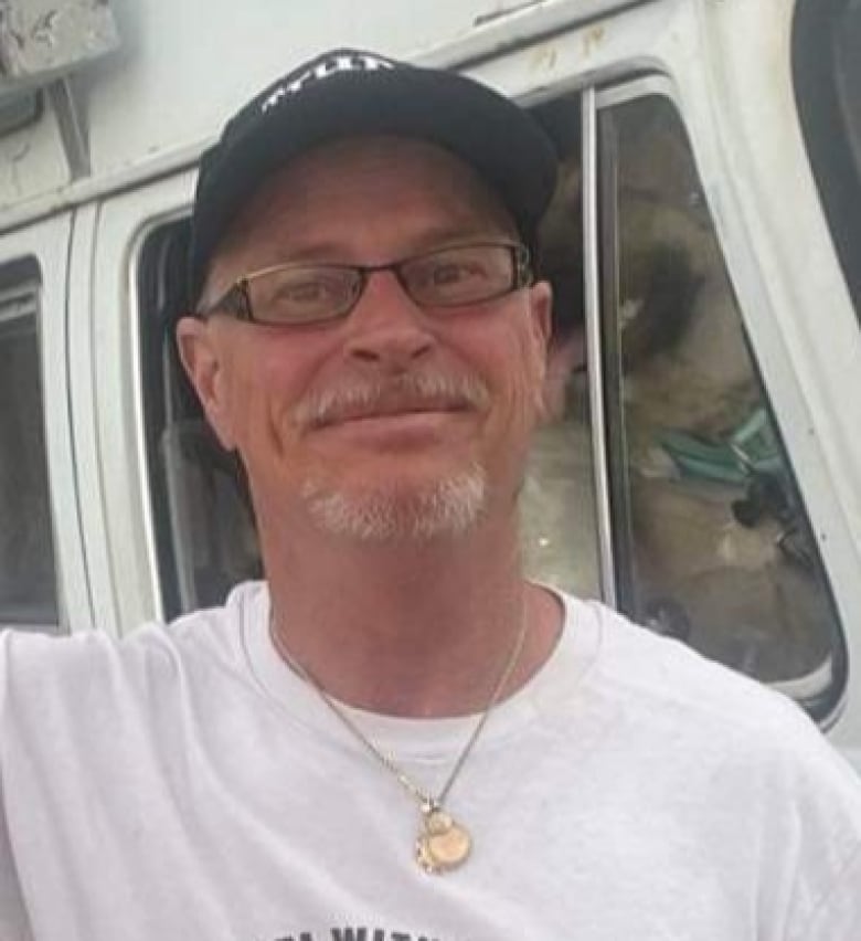 Close up of a white-haired man wearing a white shirt, a black baseball hat and glasses. 