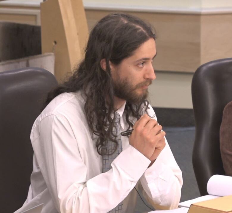 A man with long brown hair wearing a white shirt and a grey plaid tie sitting at a curved table