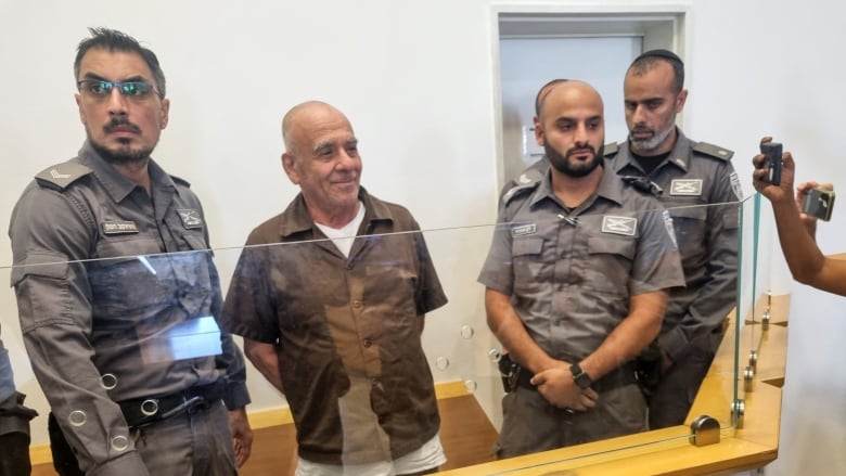 A balding, clean shaven man in a short sleeved collared shirt has his arms behind his back as he stands behind a partition in a room, surrounded by three officers in uniform.