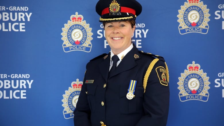 a woman officer in a black police dress uniform smiles while standing in front of a blue background with Greater Sudbury Police logos on it. 