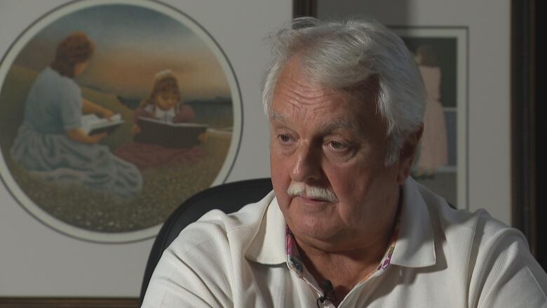 A man in a white collared shirt who has white hair and a moustache looks to the side, speaking to a reporter.