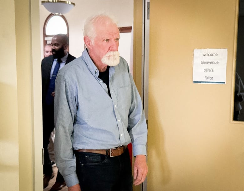 A tall thin man with thinning white hair and a white goatee wearing dark jeans and a blue denim shirt walks out of a room.