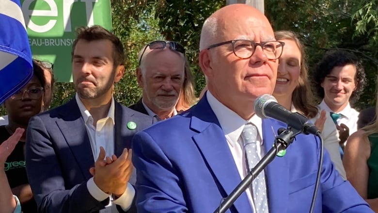 A man standing at a microphone in a blue suit with another man, in a navy suit, standing behind him.