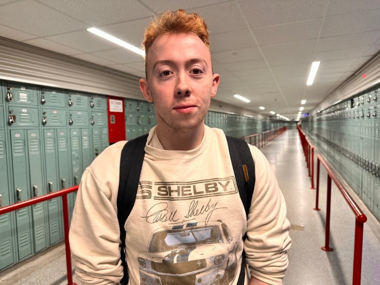 A person with short red hair and a sweatshirt with a car on it stands in a hallway lined with lockers.