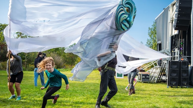 A woman carrying a giant mask-like sculpture walks along the grass. A child is running behind her.