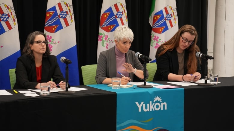 Three women sit behind a table at microphones, in front of a row of Yukon flags.