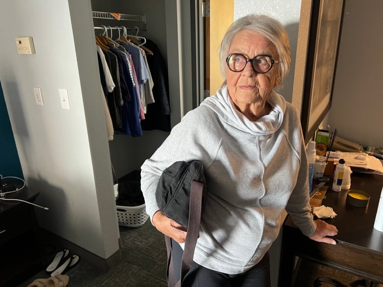 A picture of a woman with grey hard and black glasses wearing a hoodie, standing in a hotel room.