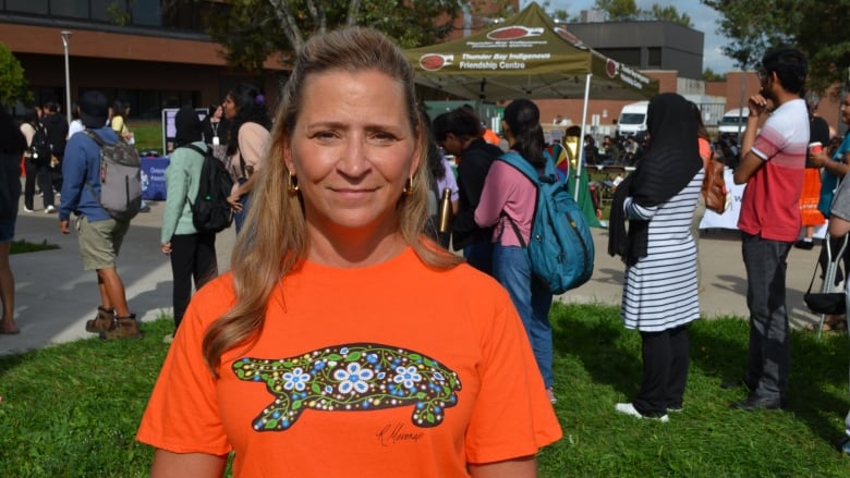 A blonde woman in an orange shirt stands in front of a tent where students are lining up.