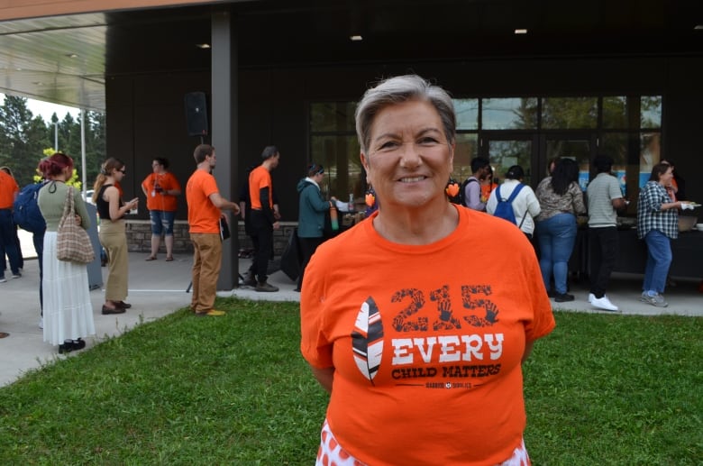 A woman with short grey hair in an orange shirts stands on the grass in front of students lined up to be served food.