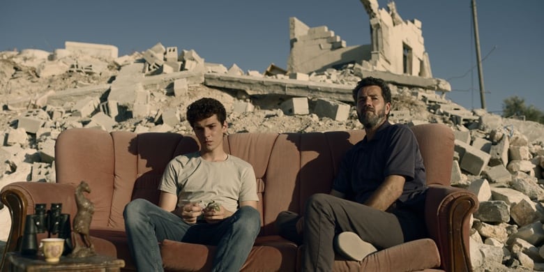 A young man and an older man sit together on couch. The couch is outdoors and behind them is a field of rubble. 