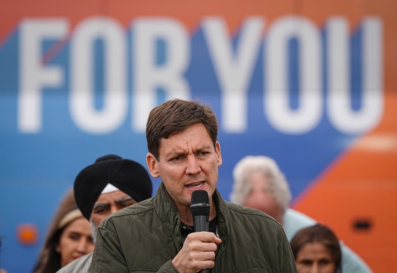 A man delivers a speech in front of supporters and a bus that reads 'For You'.