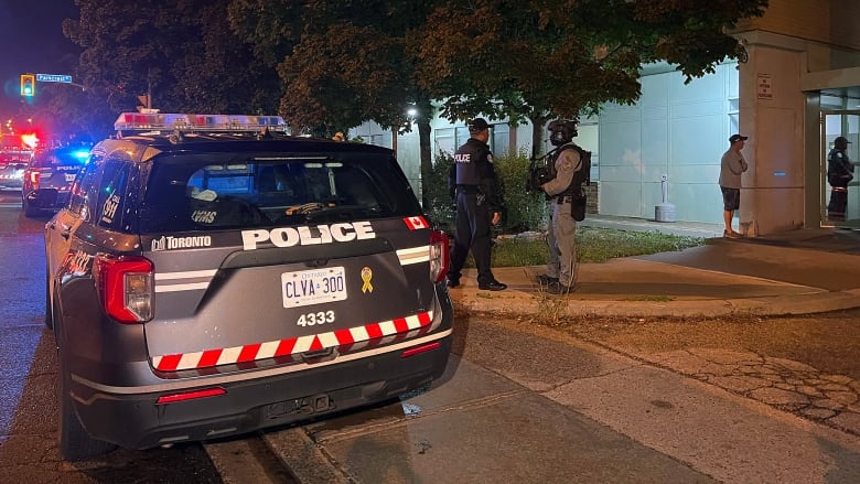 At night, a police car is parked on a city sidewalk outside a large building. 2 police officers talk to each other outside the car. One is carrying a firearm in his hands