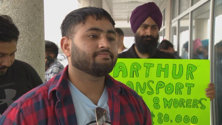 A young bearded man is visible in flannel from the chest up. He is outside a building in day time. A crowd stands around him holding signs.