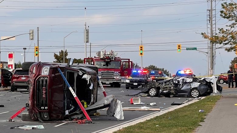 At a busy, rural intersection on a sunny day, two cars are totalled in the middle of the road. There are emergency vehicles parked in the intersection with lights flashing and police tape around the road.