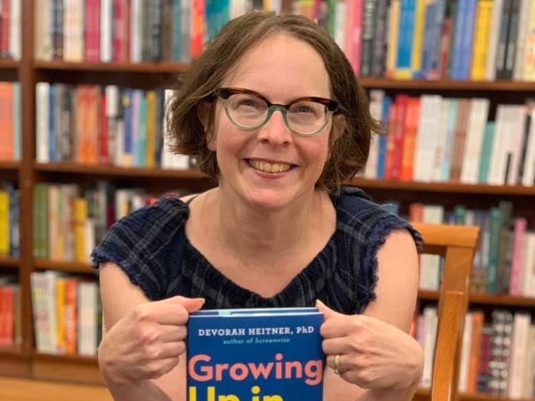 A woman with short brown hair is smiling and holding up a book with a blue cover. 