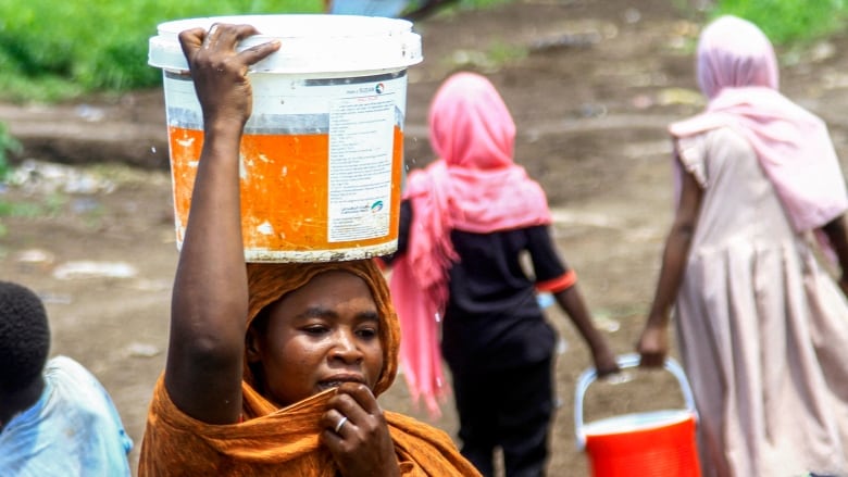 A woman walks with a plastic bucket on her head. Four other people carrying buckets can be seen in the background.