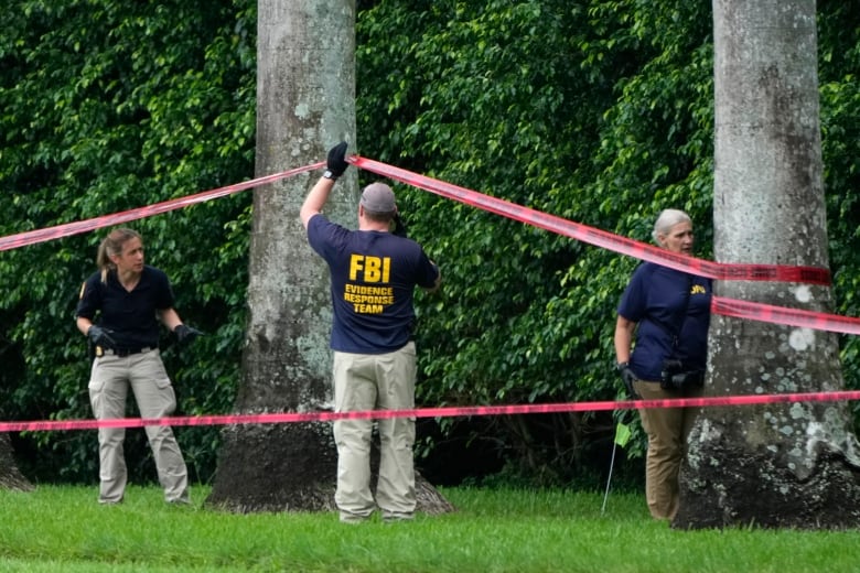 Three people in polo shorts and khaki pants, one which says FBI, are shown investigating near a treed area.