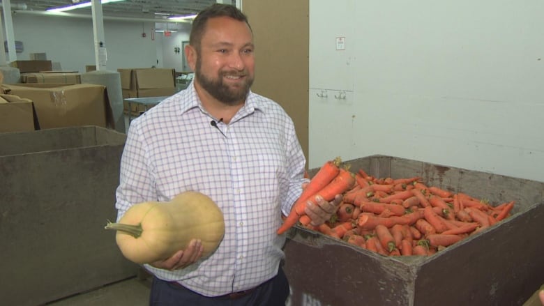 A man holding a squash and carrots in each hand smiles next to a bin full of carrots.