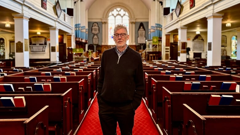 A man stands in the aisle of a church with his hands in his pocket.