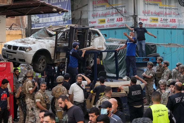 Dozens of people are shown, including military personnel in camouflage, as a damaged vehicle gets towed away in an urban setting. 