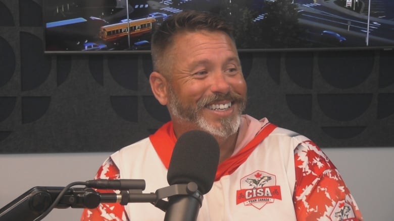 A smiling man with silver hair and a bear sits behind a microphone in a radio studio.