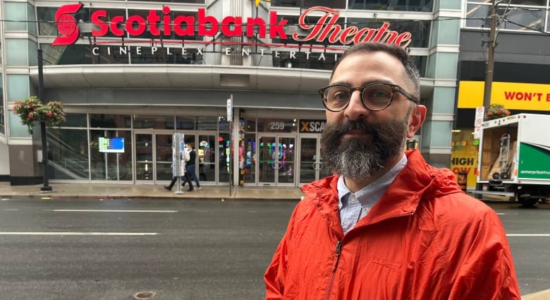 A man in an orange jacket stands in front of a theatre. 