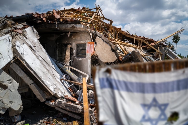 A damaged house against a blue sky. There is a tattered Israeli flag in the foreground