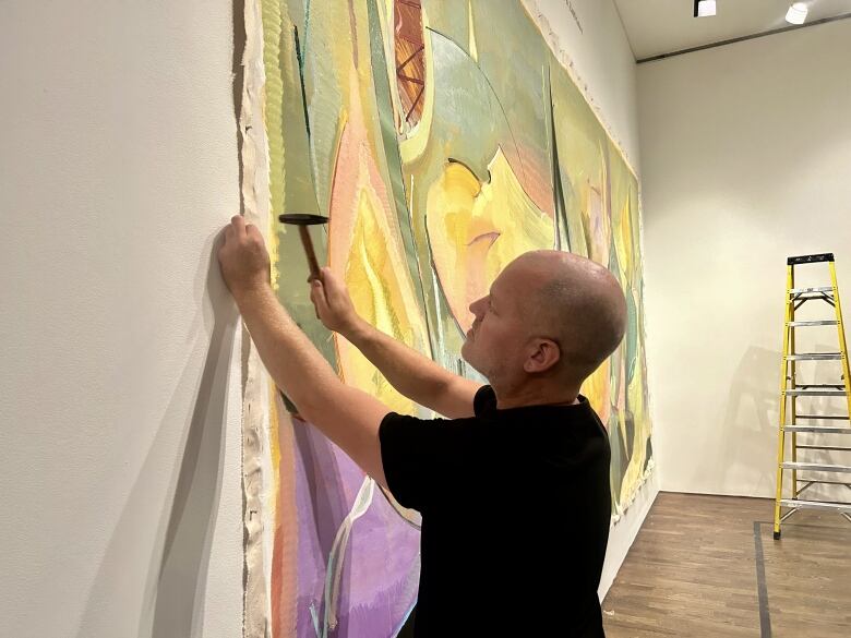 Scott Everingham standing sideways and mounting his large painting onto the wall of the AGO with a hammer Tuesday.  