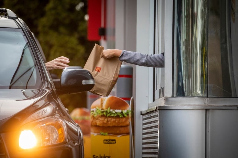 A close-up of a McDonalds employee's arm handing a fast food bag to a customer sitting in their car in a drive-through.