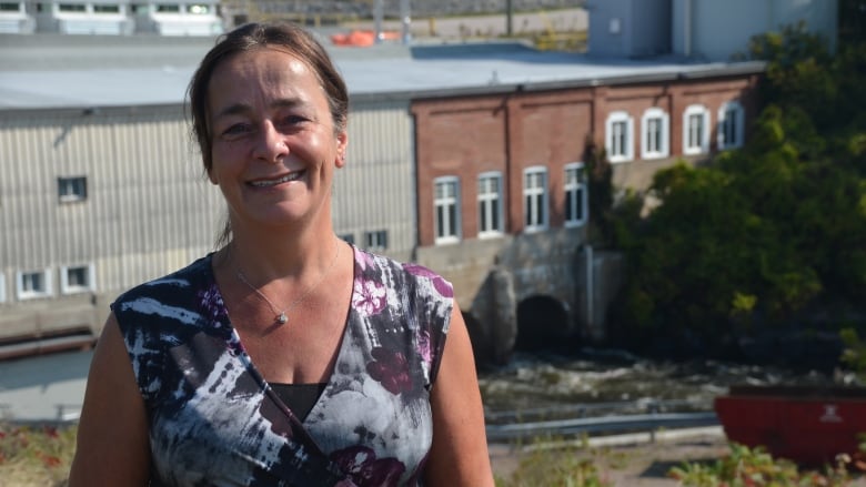 A smiling woman in a fashionable top stands in front of a hydroelectric dam 