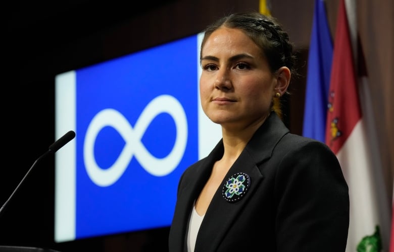 A politician is seen in front of the blue and white Mtis Nation flag.