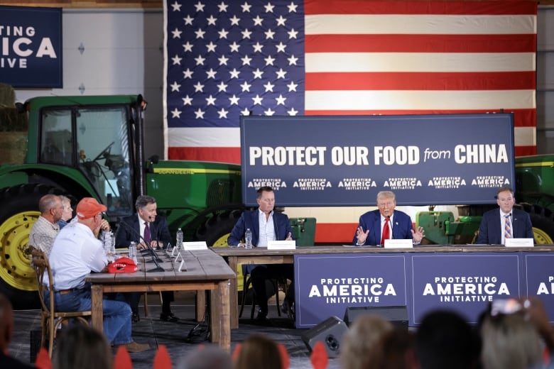 Trump at a table, in front of a green John Deere tractor and a sign that says 