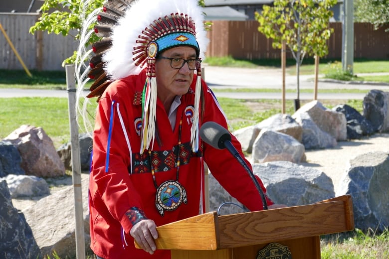 A man wearing a traditional headdress and a ribbon shirt stands at a podium.