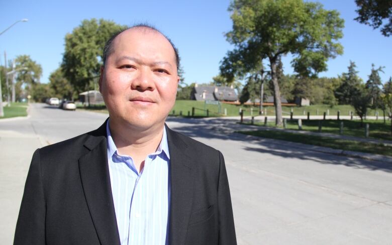 A man in a black suit with a light blue shirt underneath stands along the side of the road.