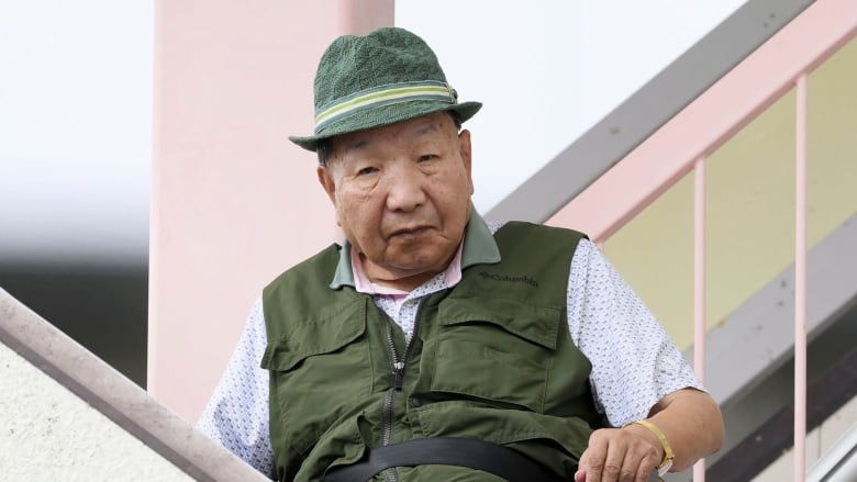 An older cleanshaven Asian man wearing a green hat and matching vest, is shown in an outdoor photo.