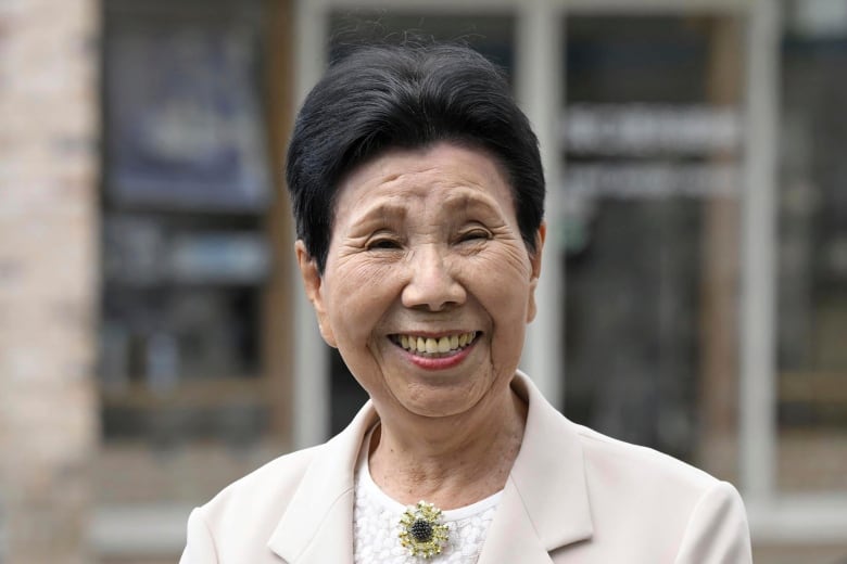 An older Asian woman wearing a collared suit jacket and a broach smiles in closeup.