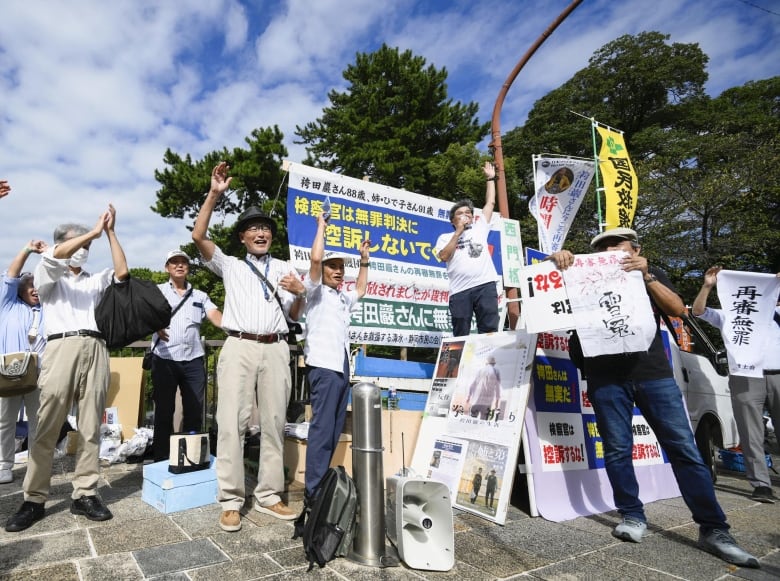 Several Asian people are shown on a sidewalk, holding signs and raising arms. One person is using a megaphone to speak.