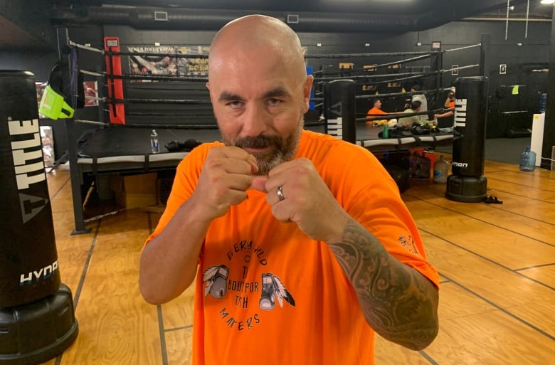 A man in an orange shirt strikes a boxer's pose in a gym.