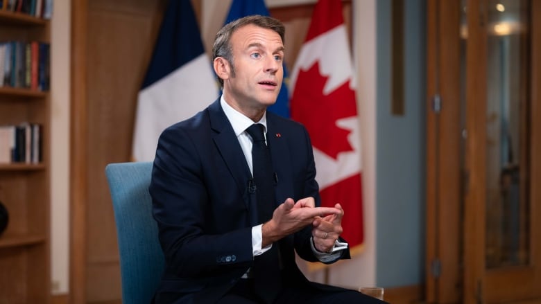 Frances President Emmanuel Macron sits in a chair wearing a blue suit as he is interviewed by CBCs Chief Political Correspondent Rosemary Barton and Radio-Canada's Parliamentary Bureau Chief Louis Blouin who are off camera. 