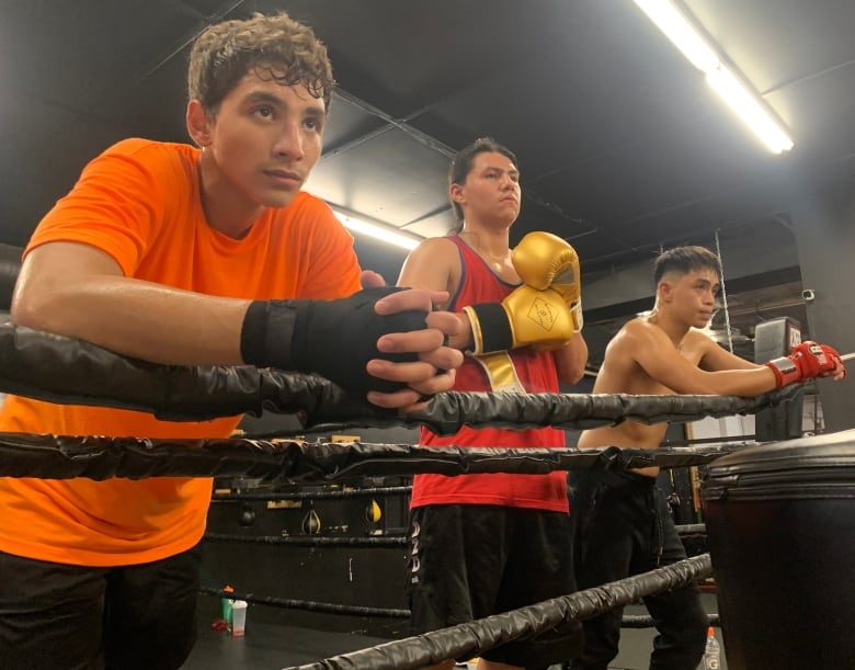 Three teenagers rest on the boxing ring ropes listening to their coach during a training session.