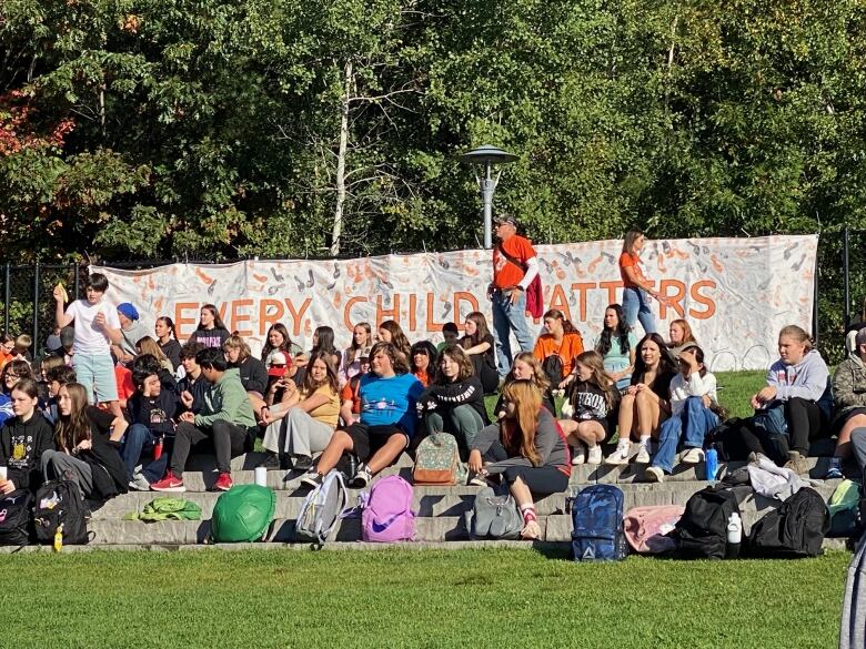 Dozens of students sit on a rock shelf in a park with a banner behind them that says 