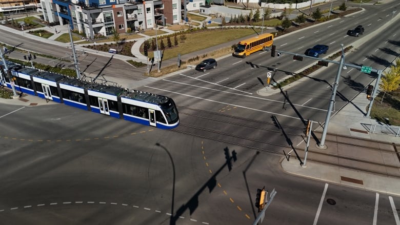 A transit train rides through a street intersection on a sunny day. Two cares and a yellow school bus are slowing down at the lights.