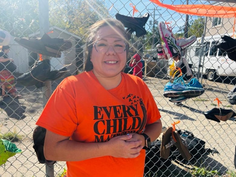 A woman in an orange shirt stands in front of a wall of shoes. 