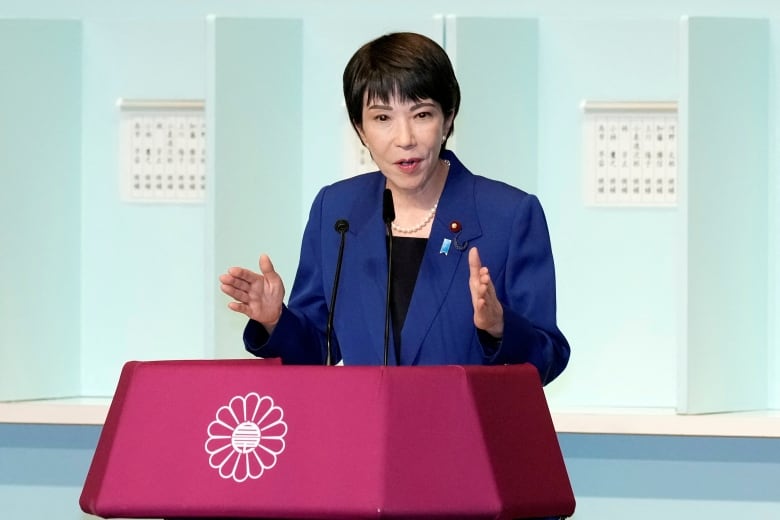 A woman with short hair wearing pearls and a blazer is shown speaking at a podium.