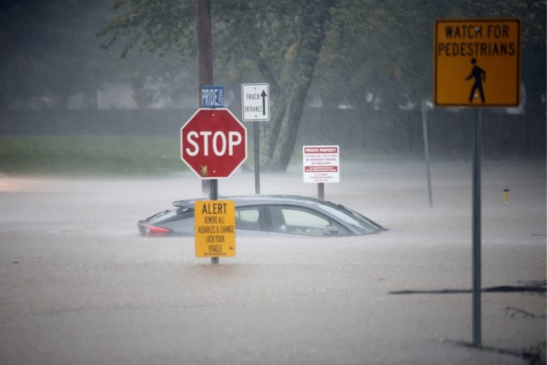 A small vehicle is barely seen above floodwater rising high on a street. A Stop sign has water well up its metal stand.