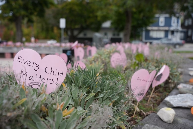 Several paper hearts stand in a flowerbed around a garden. The heart in the front reads 'Every Child Matters!'