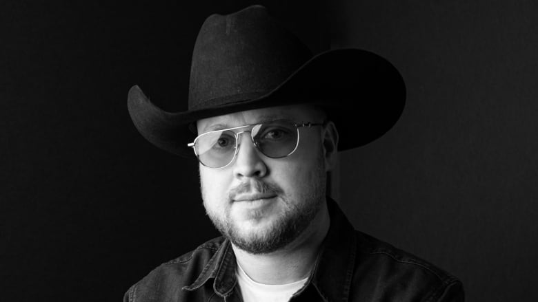 A black and white photo of a bearded man in sunglasses and a cowboy hat.