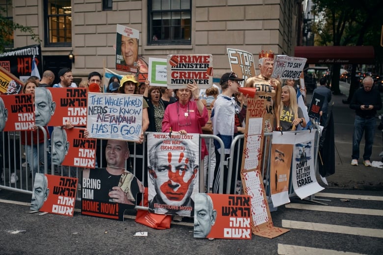 Several people hold up signs and placards on a street corner, with one wielding a loudspeaker, inside an area fenced in by metal barricades.